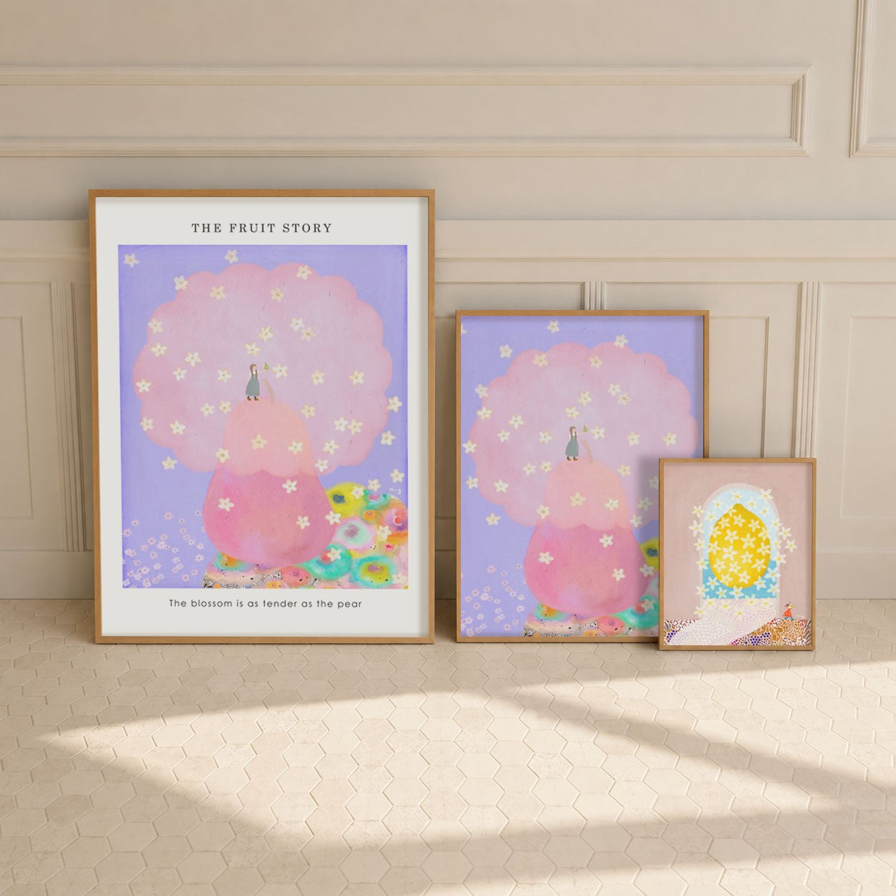 Three framed art prints with pastel illustrations are arranged against a cream paneled wall and floor. The largest print on the left reads 'THE FRUIT STORY' and shows a pink tree with small figures and flowers.