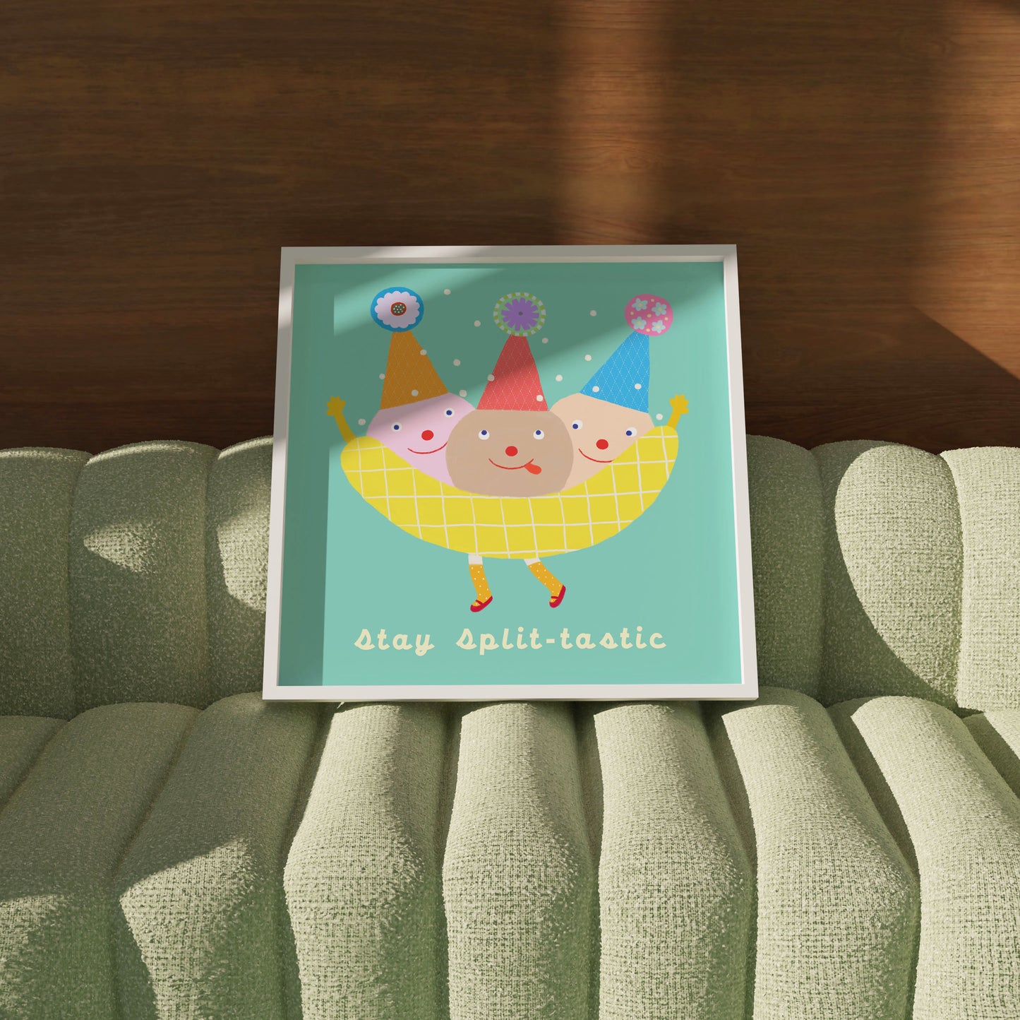 ‘Stay Split-tastic’ banana-split poster from Biliboo Shop’s haPPy scOOp Collection, resting against a sage-green sofa with a wooden backdrop.