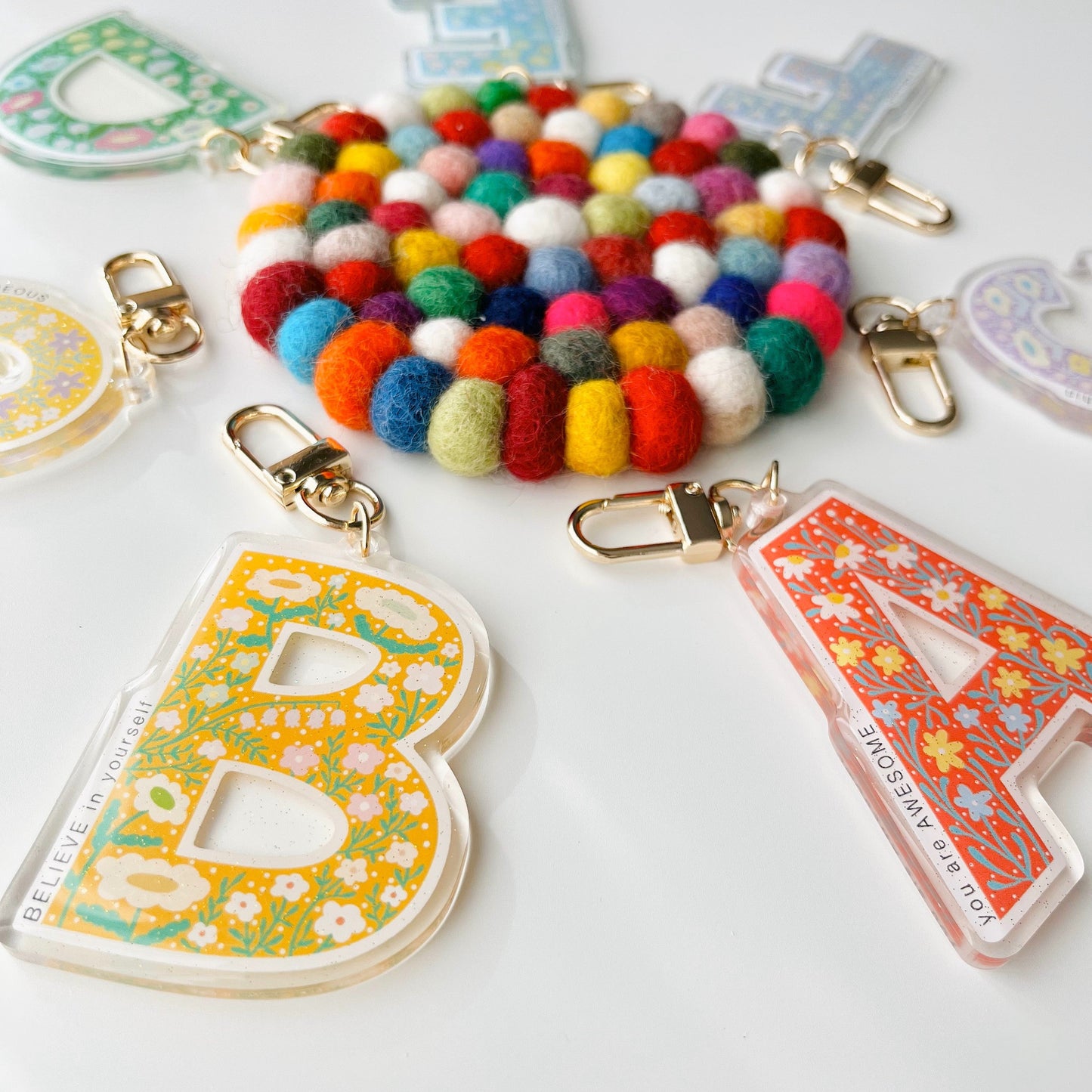 A collection of colorful letter affirmation keychains with floral patterns and glitter epoxy finish, attached to a gold-colored chain.