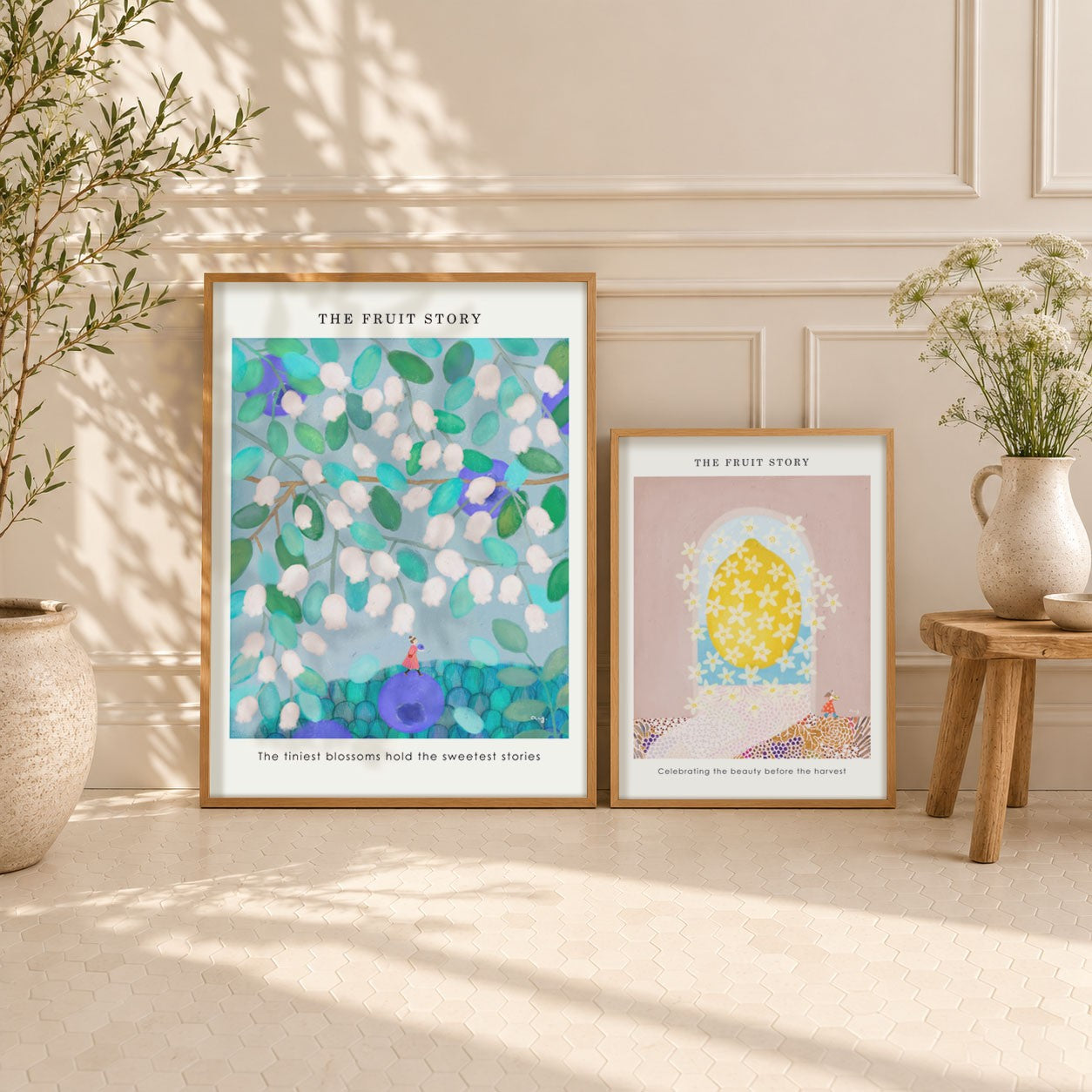 Two framed art prints titled 'THE FRUIT STORY' displayed on a light beige wall in a styled interior setting with potted plants and a small wooden stool.