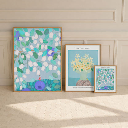 Three posters of The Fruit Story Collection are arranged on a cream tiled floor against a beige paneled wall. The largest print on the left features a whimsical blue and green floral scene with a small red figure.
