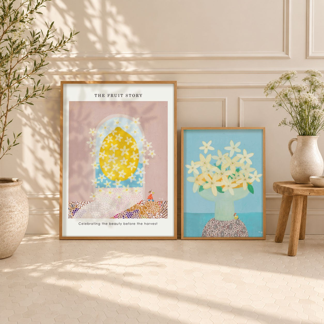 Two posters of The Fruit Story Collection displayed on a light beige floor against a cream paneled wall, styled with potted plants and a small wooden stool. 