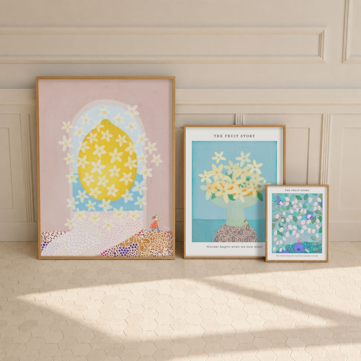 Three posters of The Fruit Story Collection are arranged on a cream-colored tiled floor against a beige paneled wall. The prints feature whimsical floral and fruit illustrations in pastel colors, including a large yellow lemon, white flowers, and blue-green botanical designs.