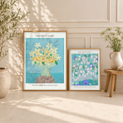 Two posters titled 'THE FRUIT STORY' displayed on a light beige wall in a styled interior setting. The larger print on the left features flowers of orange on a turquoise background, and the smaller print on the right features flowers of blueberries on a green background.