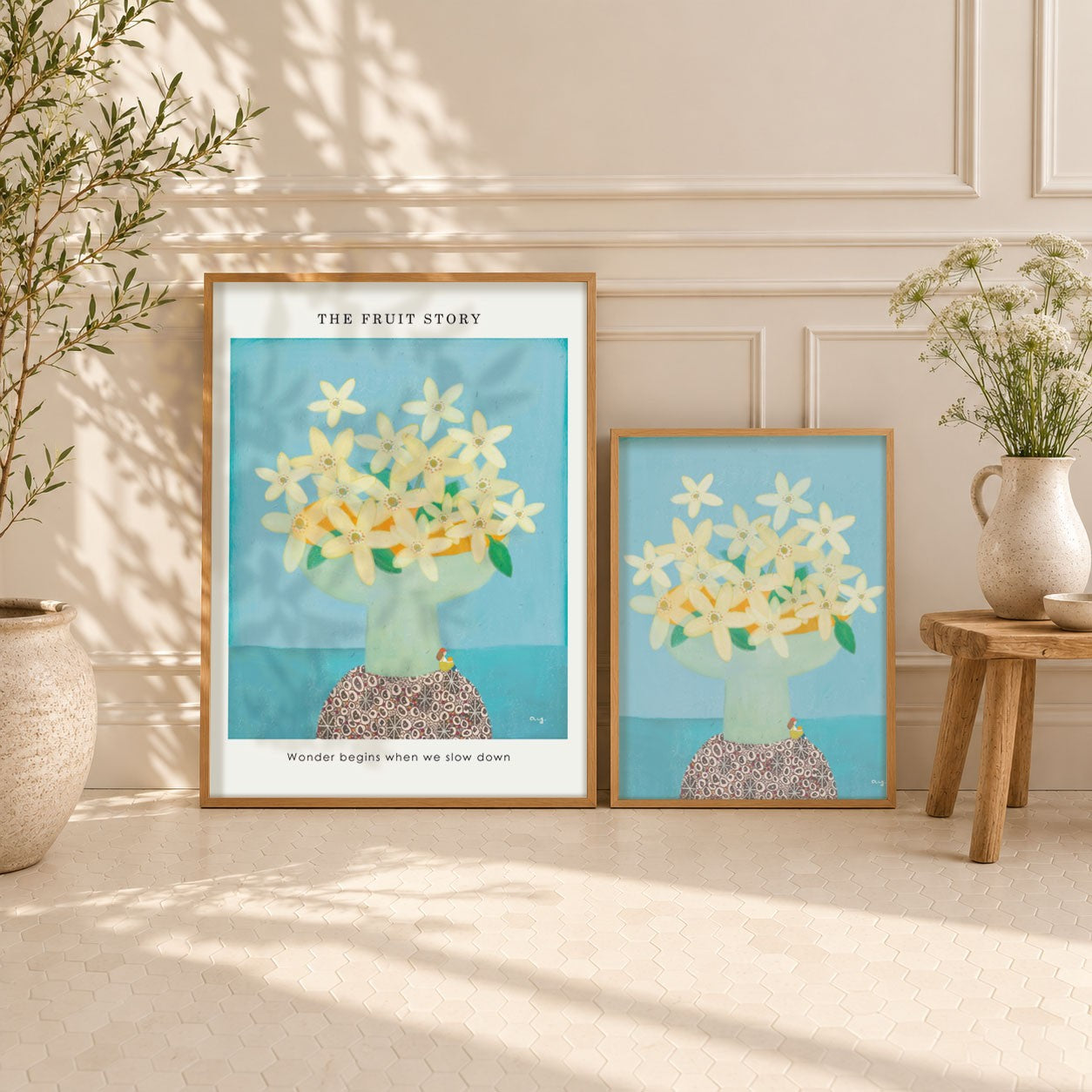 Two framed art prints featuring a stylized illustration of flowers of orange in a vase against a light blue background, styled on a cream paneled floor against a wall with potted plants.