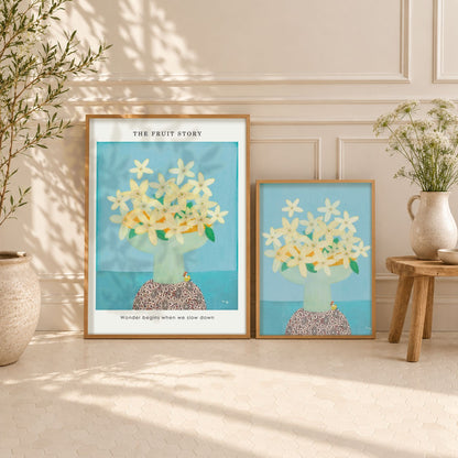 Two framed art prints featuring a stylized illustration of flowers of orange in a vase against a light blue background, styled on a cream paneled floor against a wall with potted plants.